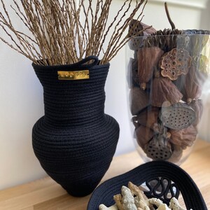 Könnte beinhalten: Schwarze Seilvase mit goldenem Etikett, gefüllt mit getrockneten Zweigen. Daneben steht eine klare Glasvase, gefüllt mit getrockneten Lotusblüten. Vor den Vasen steht eine schwarze Seil Schale gefüllt mit weißem Korallen.
