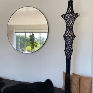 May include: A black macrame hanging with a long fringe, a round mirror reflecting a window with greenery, and two brown throw pillows on a black bench.