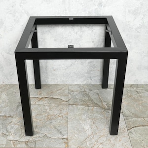 May include: A black metal table frame with a square top and four straight legs. The frame is made of square tubing and appears to be welded together. The table frame is designed to support a tabletop, and is suitable for a small table.