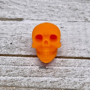 Peut inclure: Breloque en forme de cr&acirc;ne en plastique orange. Le cr&acirc;ne a un visage d&eacute;taill&eacute; avec des orbites, un nez et une bouche avec des dents.