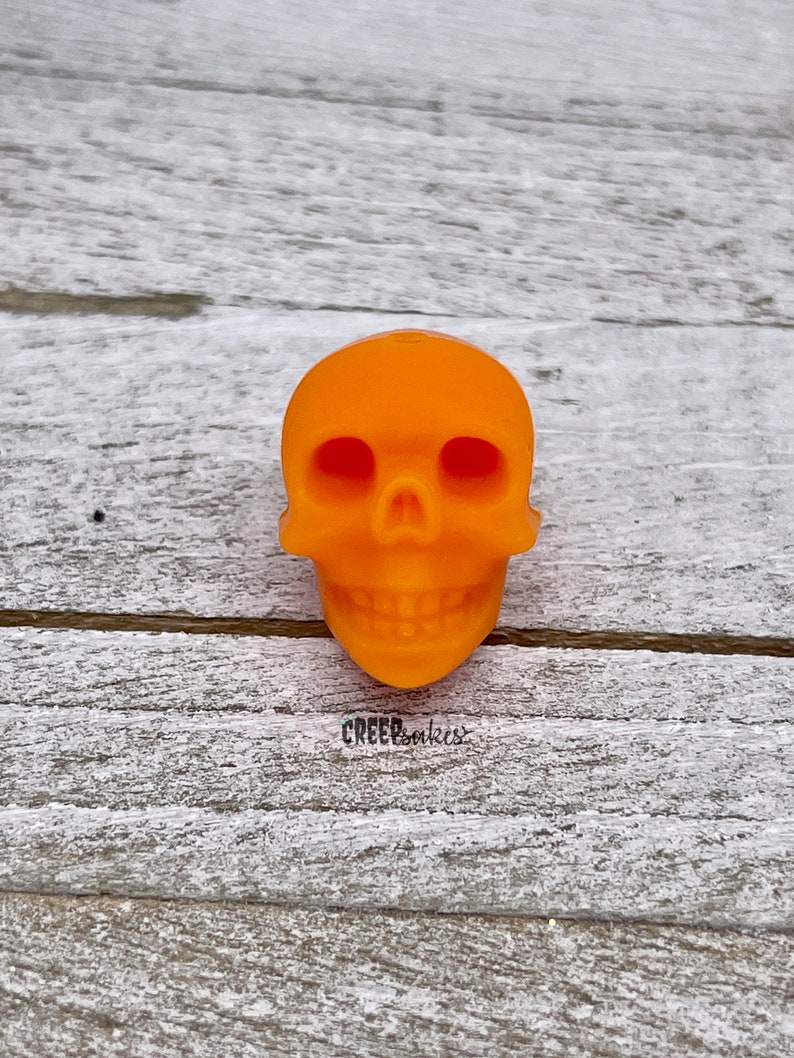 Peut inclure: Breloque en forme de cr&acirc;ne en plastique orange. Le cr&acirc;ne a un visage d&eacute;taill&eacute; avec des orbites, un nez et une bouche avec des dents.