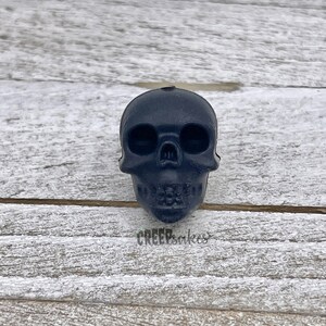 Peut inclure: Un objet en forme de cr&acirc;ne noir en r&eacute;sine. Le cr&acirc;ne a une surface lisse et repose sur une surface en bois.
