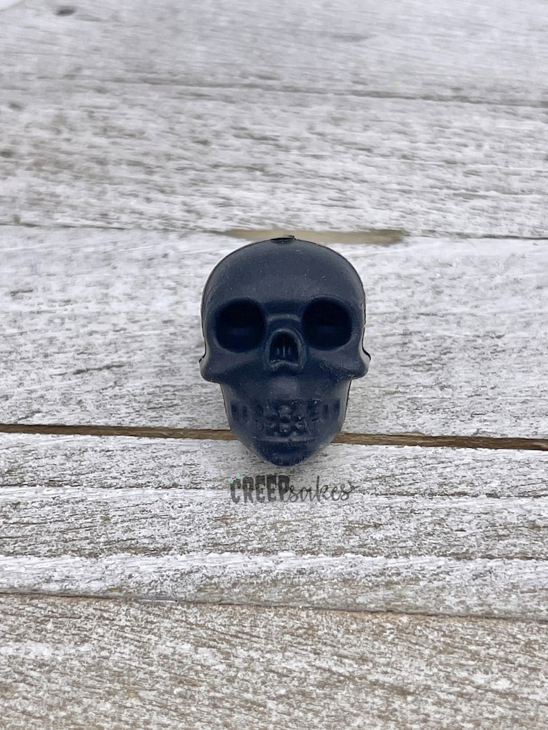 Peut inclure: Un objet en forme de cr&acirc;ne noir en r&eacute;sine. Le cr&acirc;ne a une surface lisse et repose sur une surface en bois.