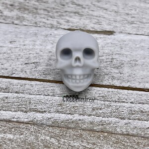 Peut inclure: Un cr&acirc;ne en c&eacute;ramique blanche avec un visage d&eacute;taill&eacute;, y compris des orbites, un nez et une bouche avec des dents. Le cr&acirc;ne est pos&eacute; sur une surface en bois clair.