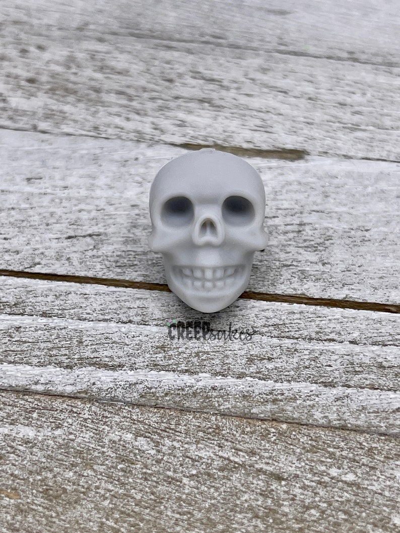 Peut inclure: Un cr&acirc;ne en c&eacute;ramique blanche avec un visage d&eacute;taill&eacute;, y compris des orbites, un nez et une bouche avec des dents. Le cr&acirc;ne est pos&eacute; sur une surface en bois clair.