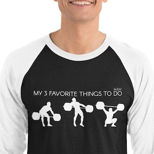 May include: A black and white baseball tee shirt with a white raglan sleeve design. The shirt features a graphic of three weightlifters with the text "MY 3 FAVORITE THINGS TO DO" above the graphic.