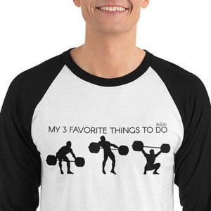 May include: A white and black baseball tee shirt with a black and white graphic of three weightlifters. The text "MY 3 FAVORITE THINGS TO DO" is printed on the shirt.