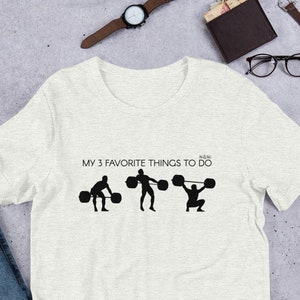 May include: A heather grey t-shirt with a black graphic of three stick figures lifting weights. The text "MY 3 FAVORITE THINGS TO DO" is printed above the graphic.