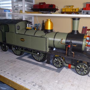 May include: A detailed model train locomotive in olive green, black, and gold. The model features intricate details, including wheels, a smokestack, and a cab. The train is displayed on a cutting mat, with other model trains visible in the background.