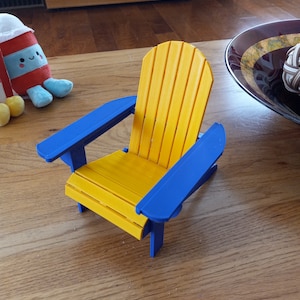 May include: A miniature yellow adirondack chair with blue armrests. The chair is made of wood and has a slatted back and seat.