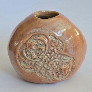 May include: A small, round, light brown ceramic vase with a textured design of a stylised creature. The vase has a small opening at the top.