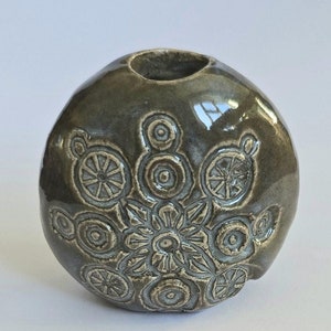 May include: A small, round, gray ceramic vase with a decorative design of circles and flowers.