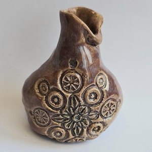May include: A small, brown ceramic vase with a unique design. The vase has a wide, round base and a narrow neck. The surface of the vase is decorated with a pattern of circles and flowers.
