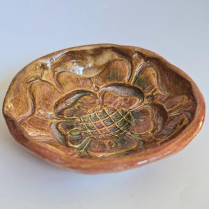 May include: A small, round ceramic dish with a floral design. The dish is glazed in shades of brown, green, and orange.