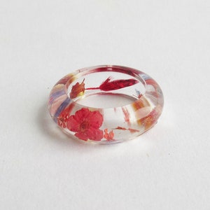 May include: A clear resin ring featuring red flowers and other botanical inclusions. The ring is circular and set against a white backdrop. The ring's diameter is approximately 2.5 cm.
