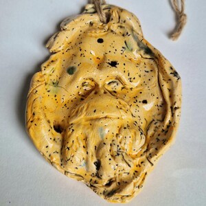 May include: A hand-sculpted clay ornament in a pale yellow hue with black speckles, featuring a moulded face with a prominent beard and moustache. The irregular, organic-shaped piece includes a hole at the top with a looped brown twine string.