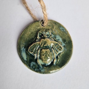 May include: A circular ceramic pendant featuring a raised bee motif. The pendant is predominantly olive green with blue highlights, and is strung on natural twine. The bee design shows detailed wings and body segments.