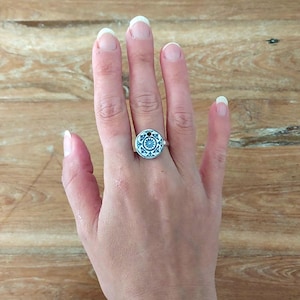 May include: A silver ring with a blue and white ceramic design. The ring is on a person's finger.