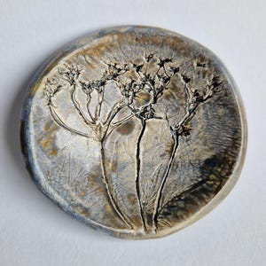 May include: A circular ceramic piece featuring a detailed tree design. The piece has a mottled colour palette of grey, brown, and blue. The tree's branches and leaves are dark, contrasting against the lighter background. The diameter is approximately 10 cm.
