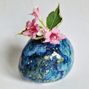 May include: A small, rounded ceramic vase with a glossy finish featuring a mottled pattern of deep blue, turquoise, and yellow speckles. The vase holds a small sprig of pink trumpet-shaped flowers with green and white variegated leaves.