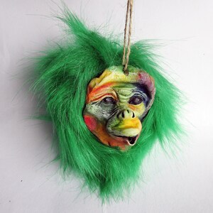 May include: A colourful monkey face ornament with a green fur wig. The monkey has a bright yellow nose and a wide, toothy smile.