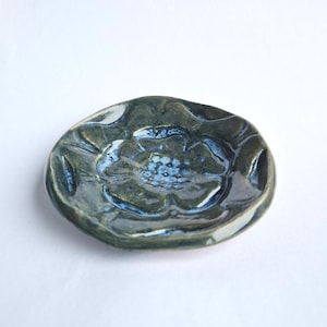 May include: A small, handmade ceramic dish with a floral design. The dish is primarily dark blue-green with lighter blue accents in the center and along the edges. The flower's petals are subtly raised, adding texture.