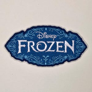 May include: An oval-shaped embroidered patch featuring a blue background with intricate light blue filigree patterns. The word Disney is embroidered in white at the top, and the word FROZEN is embroidered in large white letters below it.