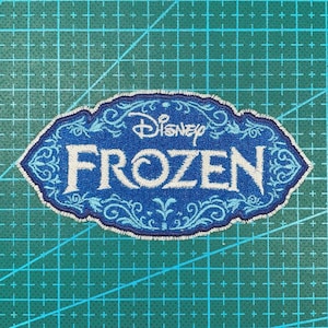 May include: An embroidered patch featuring the Disney Frozen logo in white thread on a blue background. The design is enclosed in an ornate, scalloped border with intricate light blue filigree patterns surrounding the central text.