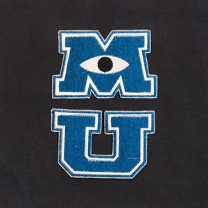 May include: Two embroidered patches featuring the letters M and U in blue with a white border. The letter M contains a white eye with a black pupil in the center. The patches are separate, with the M positioned above the U on a dark background.