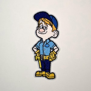 May include: An embroidered patch featuring a cartoon character wearing a blue cap, a light blue collared shirt, dark blue pants, and yellow work gloves and boots. The character has blonde hair, a pink nose, and hands placed on their hips.
