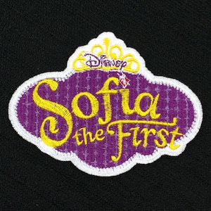 Könnte beinhalten: Lila und weiß gestickter Aufnäher mit einer Krone und dem Text "Sofia die Erste" in gelben Buchstaben.