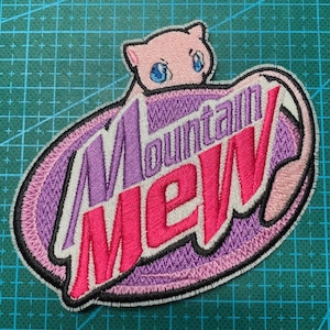 May include: An embroidered patch featuring a pink cartoon cat with blue eyes peeking out from behind a purple and pink "Mountain Dew" logo.