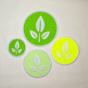 以下が含まれることがあります： 白い三つ葉の植物アイコンが刺繍された4つの円形ワッペン。ワッペンはサイズと色合いが異なり、明るいライムグリーン、淡いパステルグリーン、ネオンイエローが含まれ、それぞれ縁に白いステッチが施されています。
