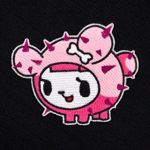 May include: Pink and white embroidered patch of a cartoon character with a cactus design. The character has a bone on its head and a smiling face.