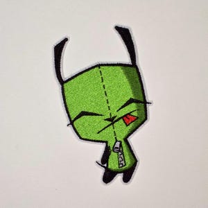 Puede incluir: Un parche bordado que muestra un personaje de color verde brillante con cabeza cuadrada, orejas negras y una pequeña lengua roja asomando. El personaje tiene un detalle de cremallera vertical en el centro del cuerpo con bordes negros.