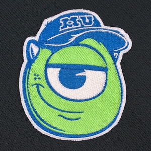 May include: An embroidered patch featuring a round, lime green character with a single large eye and a blue baseball cap with the white letters MU on the front. The patch has a white border and is set against a solid black textured fabric background.