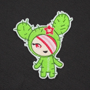 May include: An embroidered patch featuring a cartoon character wearing a green cactus suit with two large ears, a red star accent, and a pink and white striped headband. The character has a smiling face, black eyes, and a green body with white stitching.