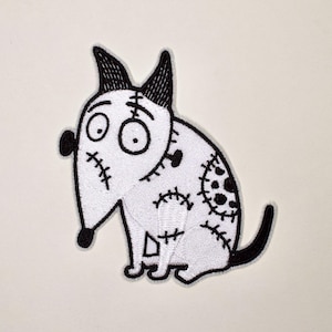 May include: An embroidered iron-on patch featuring a stylized white dog with black outlines. The dog has large circular eyes, pointed ears, and various stitched-looking markings on its body, including a circular patch on its side and a tail.