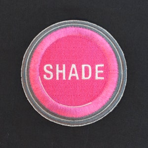 May include: A circular embroidered patch featuring the word "SHADE" in white capital letters. The patch has a vibrant pink centre, a grey border, and a thin white outline. The patch is set against a black background.
