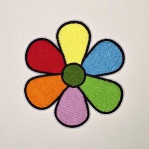 May include: A flower-shaped embroidered patch featuring six rounded petals in yellow, blue, green, light purple, orange, and red, surrounding a solid green circular center. Each petal and the center are outlined with a thick black border.