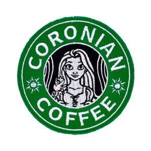 May include: A circular embroidered patch featuring a green border with the white text CORONIAN COFFEE and two sun icons. The center displays a black circle with a white embroidered illustration of a long-haired character holding a small lizard.
