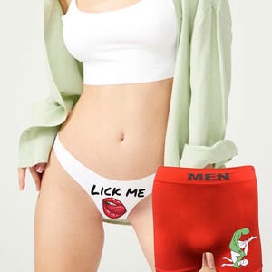 May include: A white thong featuring the black text LICK ME above a red graphic of a mouth with a tongue, and a pair of red boxer briefs with a black waistband labeled MEN and a graphic of two figures in a green and white color scheme.