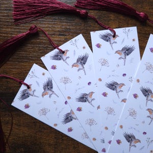 Peut inclure: Ensemble de quatre marque-pages blancs ornés d'un motif répété d'oiseaux, de fleurs et de feuilles. Chaque marque-page est doté d'un pompon bordeaux. Les marque-pages sont disposés sur une surface en bois.