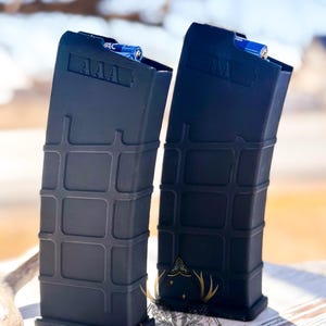 May include: Two black rectangular magazines with a grid pattern are displayed. Each magazine has a blue battery inside. The top of each magazine has the letters "AAA" printed on it. The magazines are on a wooden surface.