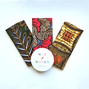 Peut inclure: Vue aérienne de trois morceaux de tissu pliés avec des motifs colorés de wax africain. Un autocollant rond blanc avec le texte "WAX & WOVEN" est au centre. Les tissus présentent des teintes vertes, rouges et jaunes.