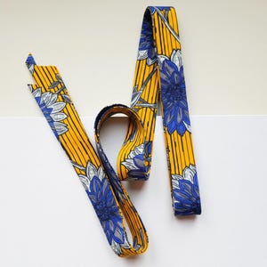 Peut inclure: Une longue bande de tissu étroite présentant un fond jaune vif avec des lignes noires verticales et un motif floral répétitif dans des tons de bleu, blanc et gris. Le textile est plié en boucle, mettant en valeur l'imprimé botanique coloré.