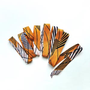 Pode incluir: Uma coleção de tiras de tecido estampadas em tons de amarelo, preto, branco e rosa. As tiras estão dispostas sobre um fundo branco, mostrando vários desenhos geométricos e abstratos. O tecido parece estar dobrado e disposto em forma de leque.