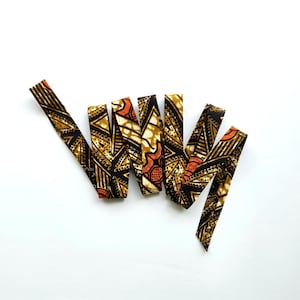 May include: Several fabric strips with a colourful African print. The strips display a repeating pattern of black, brown, orange, and cream geometric shapes. The strips are arranged on a white background.