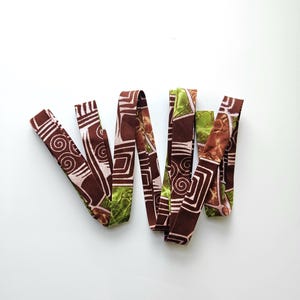 Puede incluir: Varias tiras de tela con patrones geométricos en marrón, verde y blanco se disponen sobre un fondo blanco. Las tiras presentan una variedad de diseños, incluyendo espirales y formas angulares. La tela parece ser un adorno decorativo.