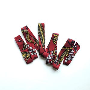 Puede incluir: Varias tiras de tela roja con un patrón repetido de diseños amarillos y negros y círculos blancos. Las tiras están dispuestas sobre un fondo blanco. La tela parece ser un textil decorativo.